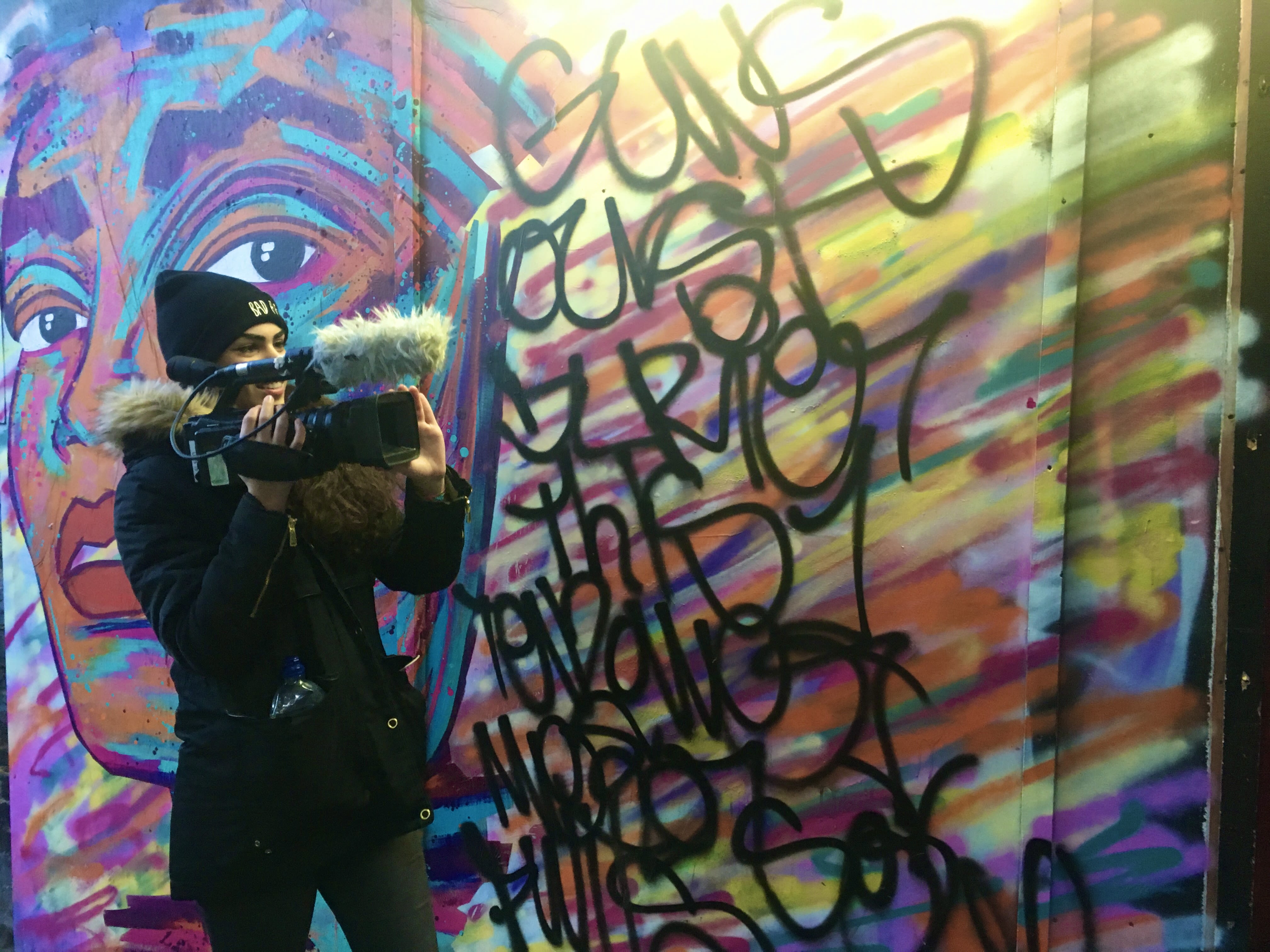 Ela holding a camera, filming a street artist painting a wall