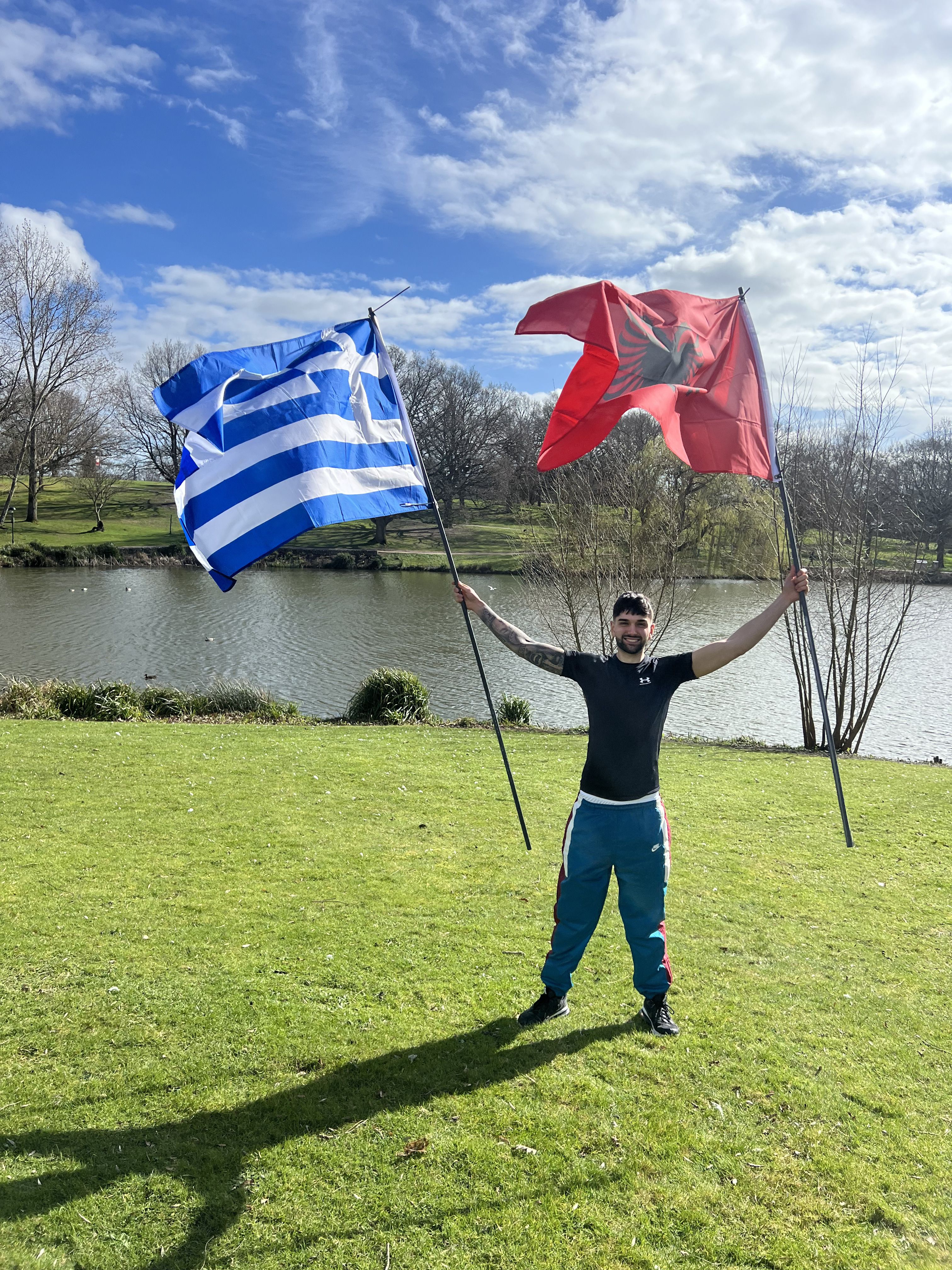 Michael stood in front of the lake at Colchester Campus holding the Greek and Albanian flags